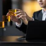 Businessman in suit carefully stacking amber-colored glass cubes at a desk with a laptop.