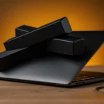 Laptop with black headphones, hourglass, and yellow speaker on wooden desk against orange background.