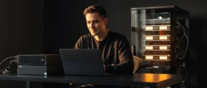 Man working on laptop at desk with computer server and networking equipment in dark studio setting.