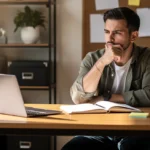 Man in olive shirt sitting at wooden desk with laptop and notebook, hand to chin in thoughtful pose.