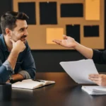 Two professional men in business meeting discussing documents at table with modern office background