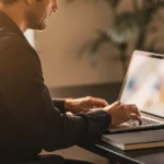 Man working at laptop at desk with coffee mug and notebook in warm office lighting.