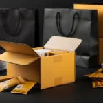 Open cardboard box surrounded by packaging materials, shipping envelopes, glasses, and black shopping bags on dark surface.