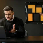 Man in black sweater gestures while looking at laptop, with two women pointing at a board with yellow and black squares behin