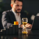 Hourglass and ascending wooden blocks on desk with businessman working at laptop in background.