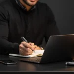 Man in black hoodie writing in notebook at desk with laptop and product containers with black and gold packaging.