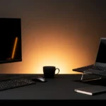 Dark desk setup with monitor, laptop, keyboard, mouse, coffee mug, notebook, and headphones lit by warm backlighting.