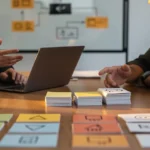 Two people discussing a laptop at a wooden table with colorful sticky notes and notecards arranged in front of them.