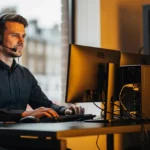 Man wearing headset at desk with computer monitors and server equipment in an office overlooking a city street.