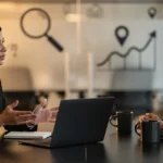 Two business professionals in formal attire discuss work at a conference table with a laptop and coffee mugs.