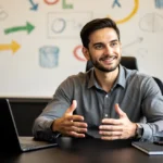 Man in gray shirt gesturing while speaking at desk with laptop, whiteboard with colorful diagrams in background.