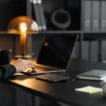 Person working at desk with laptop, coffee mug, crumpled papers, and sticky notes in a dark office environment.