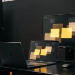 Server rack with illuminated indicator lights next to a laptop and monitor displaying yellow and tan colored squares.
