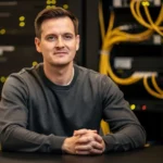 Man in gray shirt seated at table in server room with yellow network cables in background.