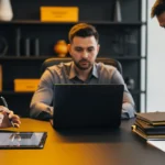Three businessmen review documents and a laptop at a conference table in an office.