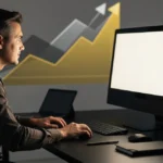 Man at desk working on computer with upward trending chart graphic displayed on wall behind him.