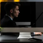 Man in business attire sits at desk with hourglass, plant, and computer monitor against blurred city lights background.