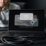 Laptop displaying design tool icons with cables and peripherals on desk, person in background.