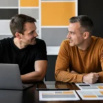 Two men in casual shirts discuss work at a desk with a laptop and design mockups in front of a geometric wall display.