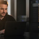Man in dark hoodie working at laptop in server room with illuminated equipment racks.