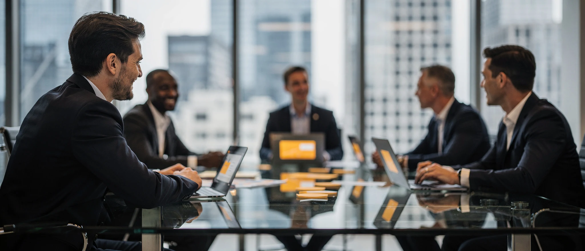 Business professionals in formal attire seated around a conference table with laptops, overlooking a city skyline.