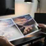 Person holding an open photo book with landscape photographs at a desk with computer monitors.