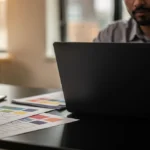 Man in grey shirt working at laptop in high-rise office with colour swatches and smartphone on desk.