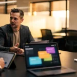 Three men in business attire discuss a project around a table with tablets and laptops displaying coloured interface designs.