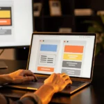 Designer working on website layouts displayed across a monitor, laptop, and tablet showing UI mockups with colored sections.