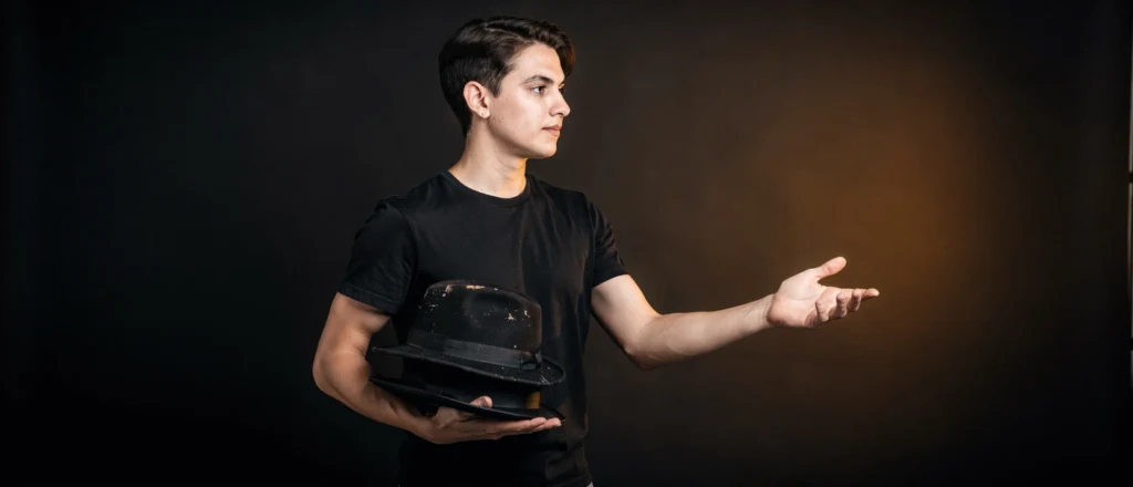 Young man in black t-shirt holding a worn black hat, gesturing with one hand against a dark background.