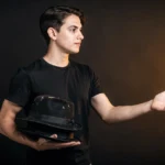 Young man in black t-shirt holding a worn black hat, gesturing with one hand against a dark background.