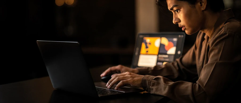 Man working at a laptop at night with a tablet displaying design software in the background.