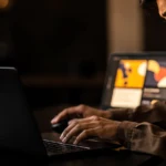 Man working at a laptop at night with a tablet displaying design software in the background.