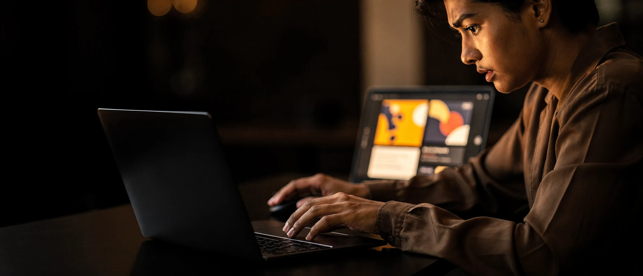 Man working at a laptop at night with a tablet displaying design software in the background.