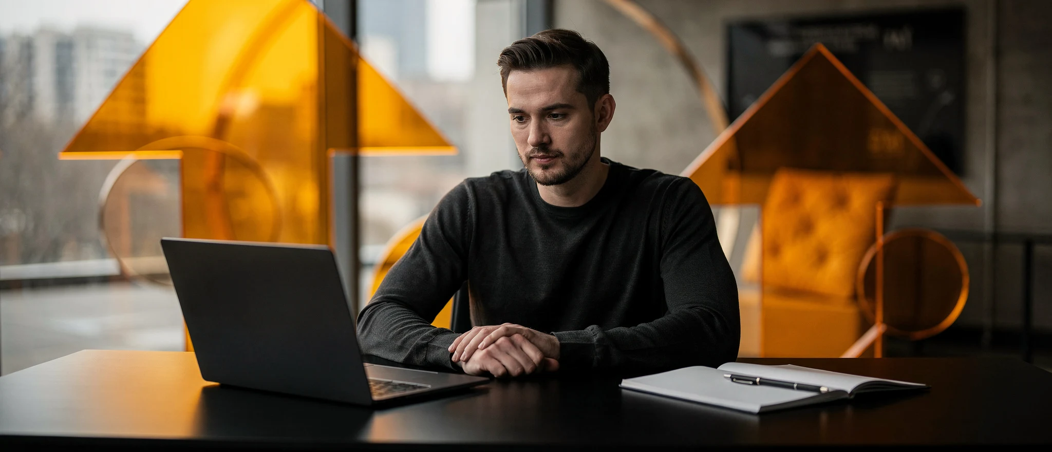 Man in black shirt working at laptop at desk with yellow geometric sculptures and city skyline visible through window behind